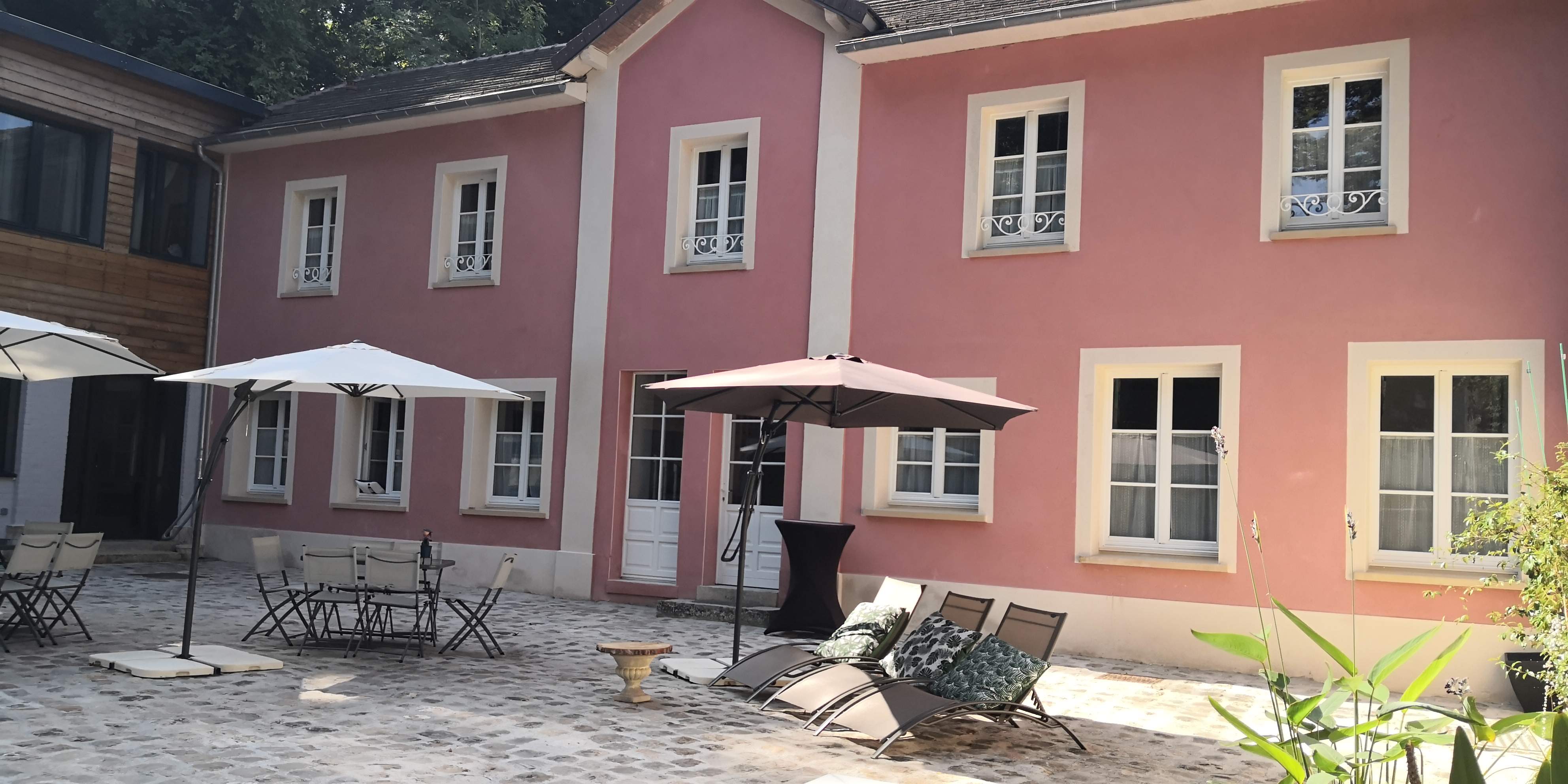 Façade rose de la maison d'hôtes vue depuis la cour pavée, mobilier de terrasse et parasols.