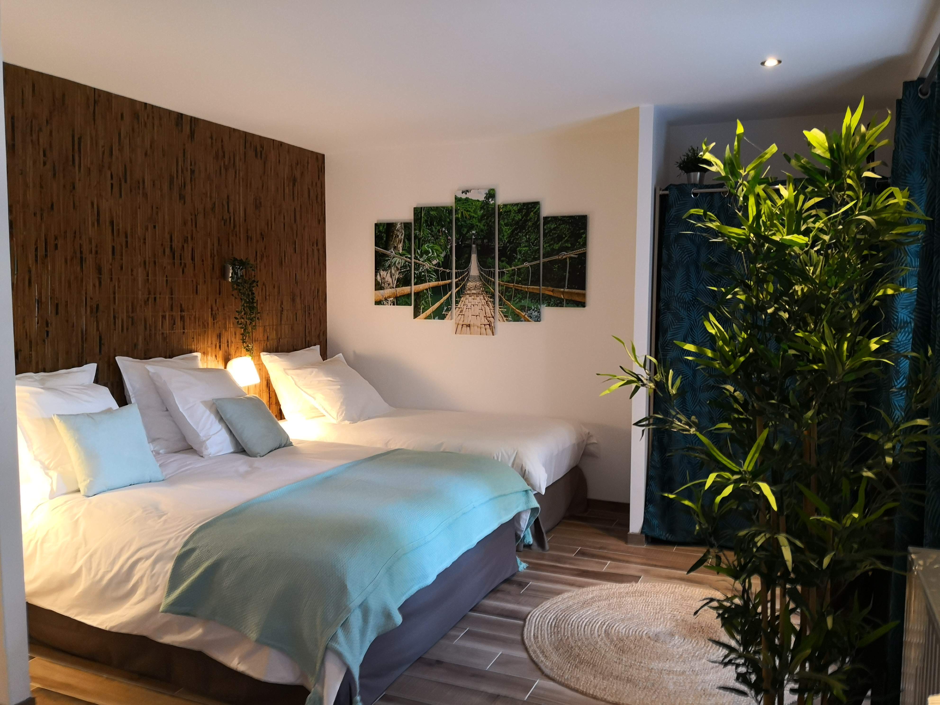 Chambre avec deux lits à tête de lit en bambou et jetés turquoise, toile représentant un pont de bambou dans la jungle au mur.