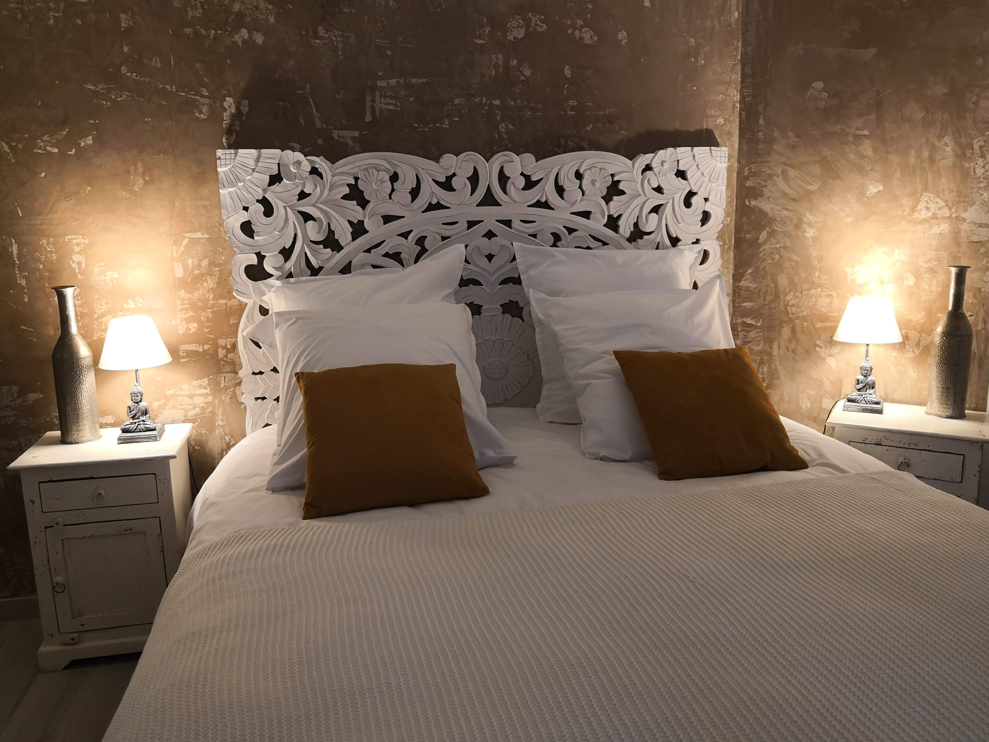 Lit avec tête de lit en bois sculpté et blanchi, coussins blancs et velours moutarde, mur en enduit patiné bronze et lampes à base de bouddha argentée de part et d'autre.