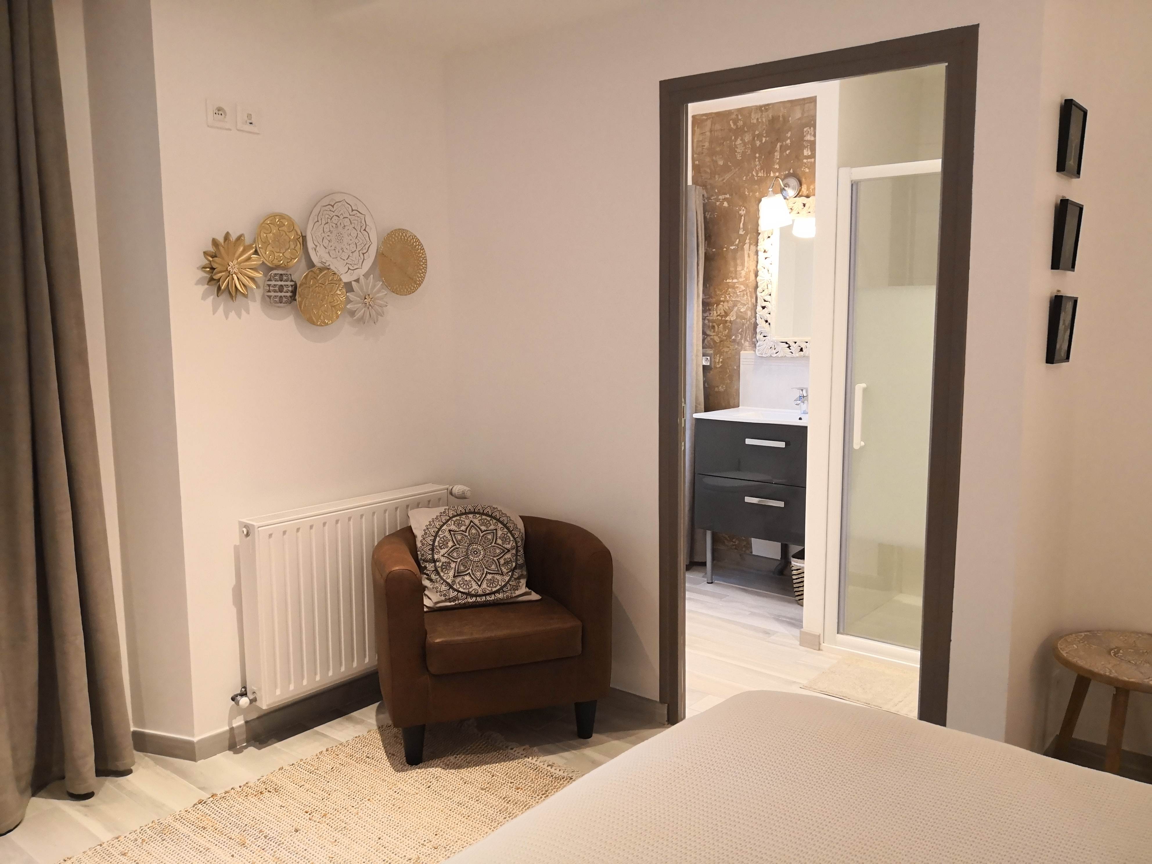 Coin de chambre avec fauteuil en cuir brun, coussin motif mandala, plats décoratifs dorés au mur et vue sur la salle de bain attenante par une porte ouverte.