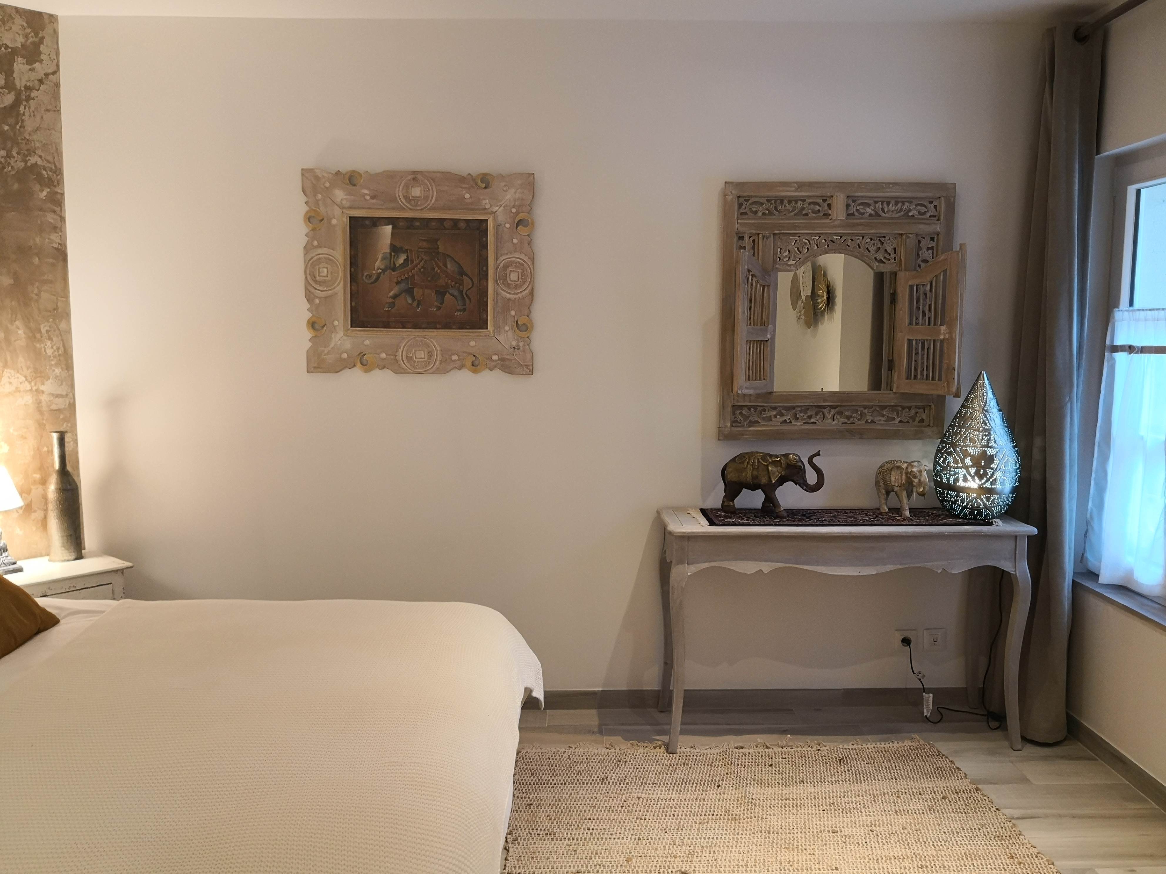 Chambre avec console en bois patiné, statuettes d'éléphants en bronze, lampe marocaine ajourée et miroir en bois sculpté à volets, près d'une fenêtre voilée.