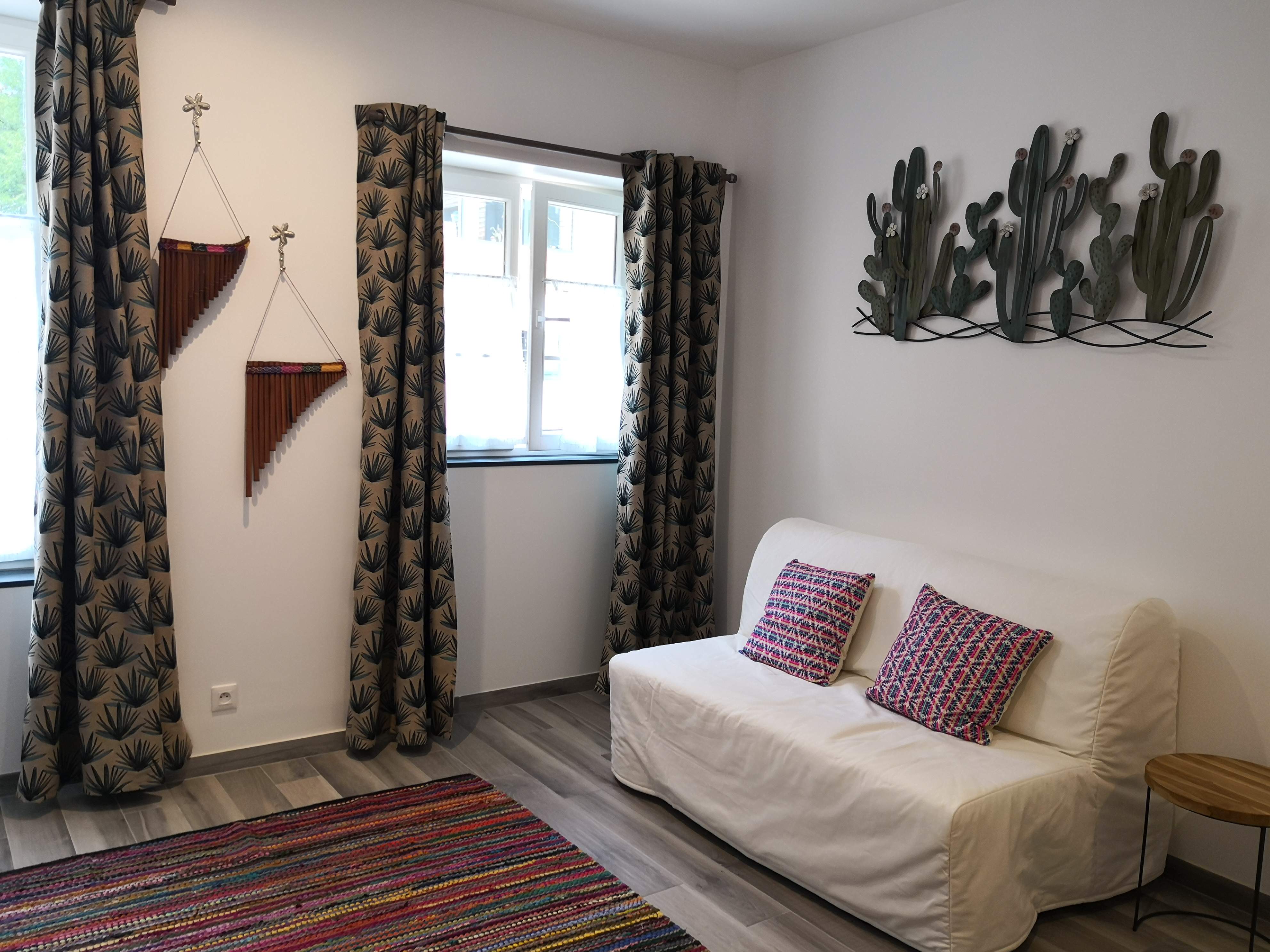 Coin salon d'une chambre avec canapé blanc, coussins à motifs, tapis tissé multicolore et décoration murale de cactus en métal.