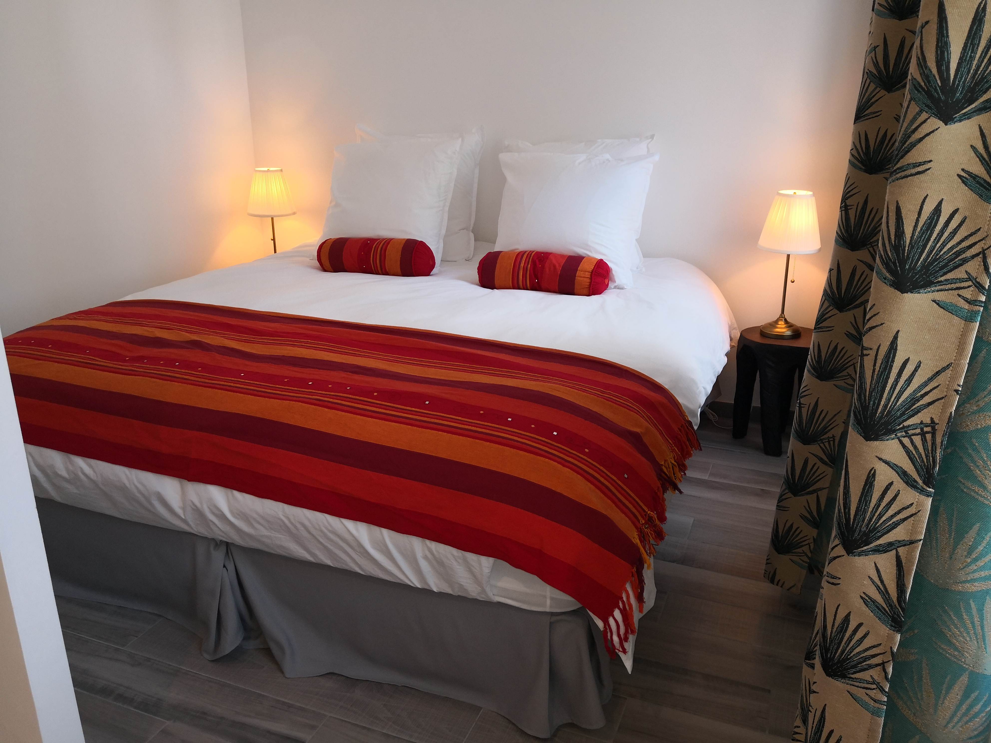 Lit double avec couvre-lit rayé rouge, orange et bordeaux à pampilles et oreillers blancs, rideaux à motif de feuilles vert foncé, dans une chambre d'hôtes.