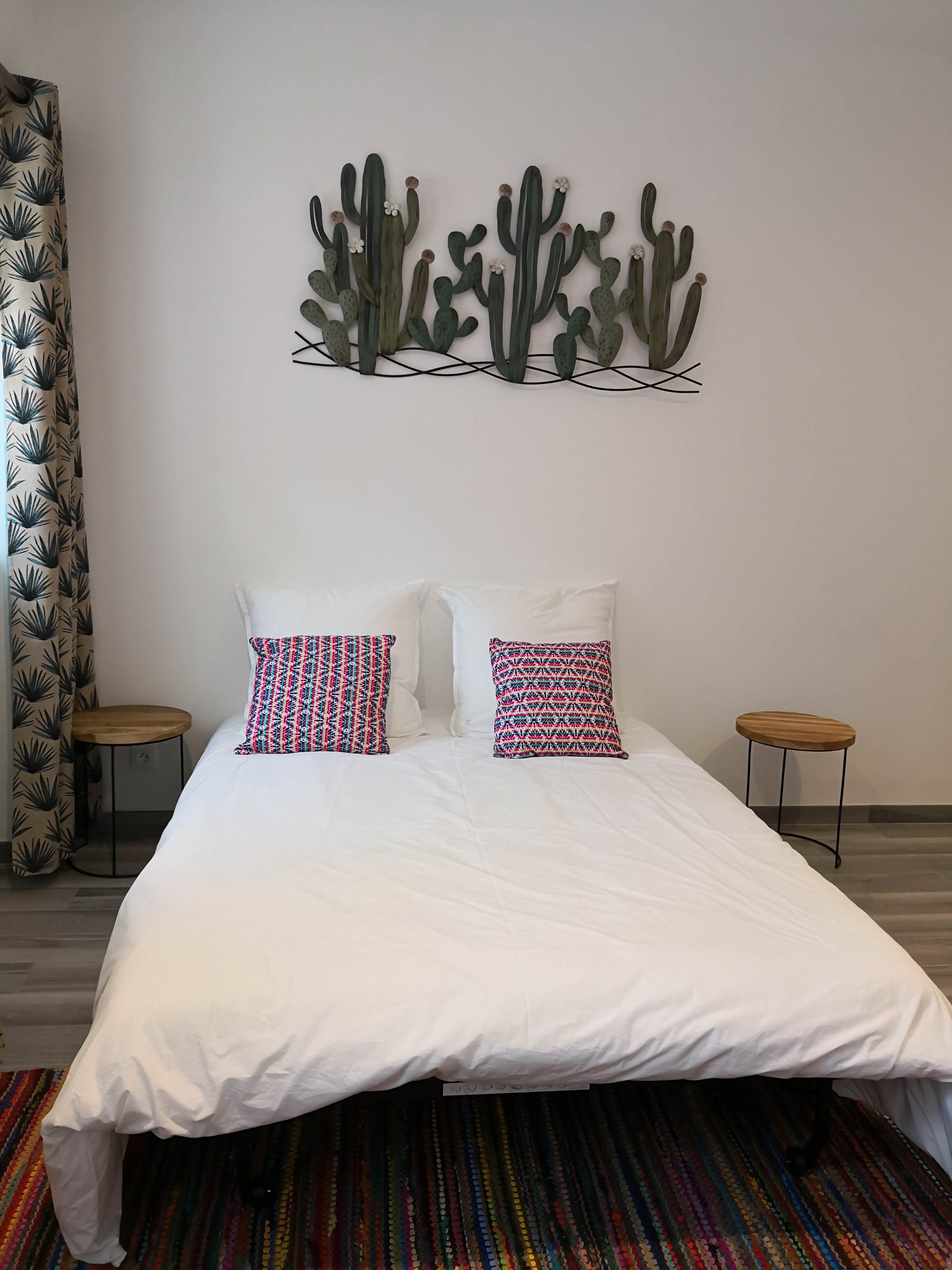 Chambre avec lit double aux draps blancs, coussins à motif géométrique rose et bleu marine, décoration murale en cactus métallique, rideau à motif de feuilles de palmier, deux tables de chevet rondes en bois clair.