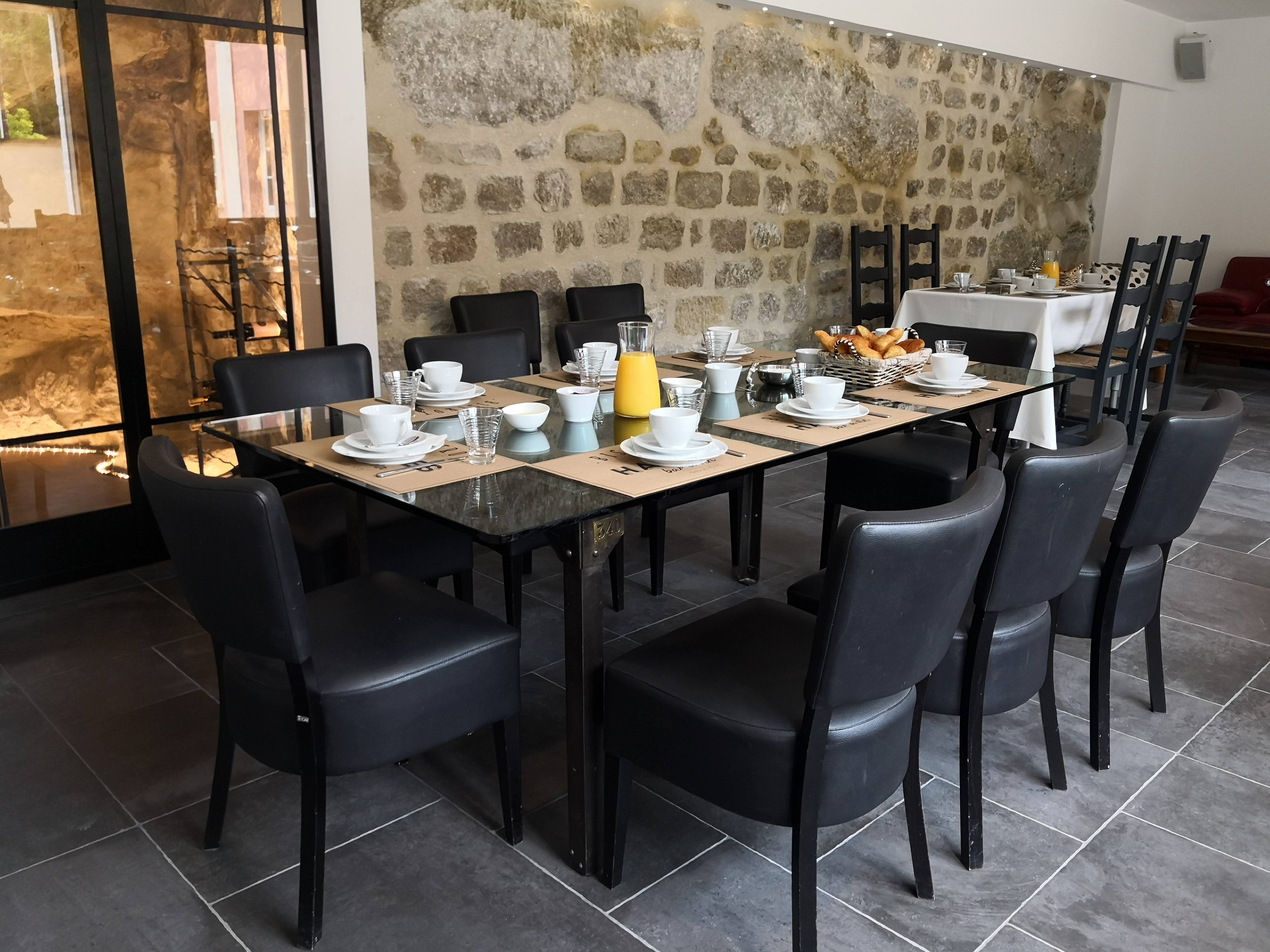 Table du petit-déjeuner dressée avec viennoiseries, confitures et jus d'orange, devant un mur en pierre apparente.