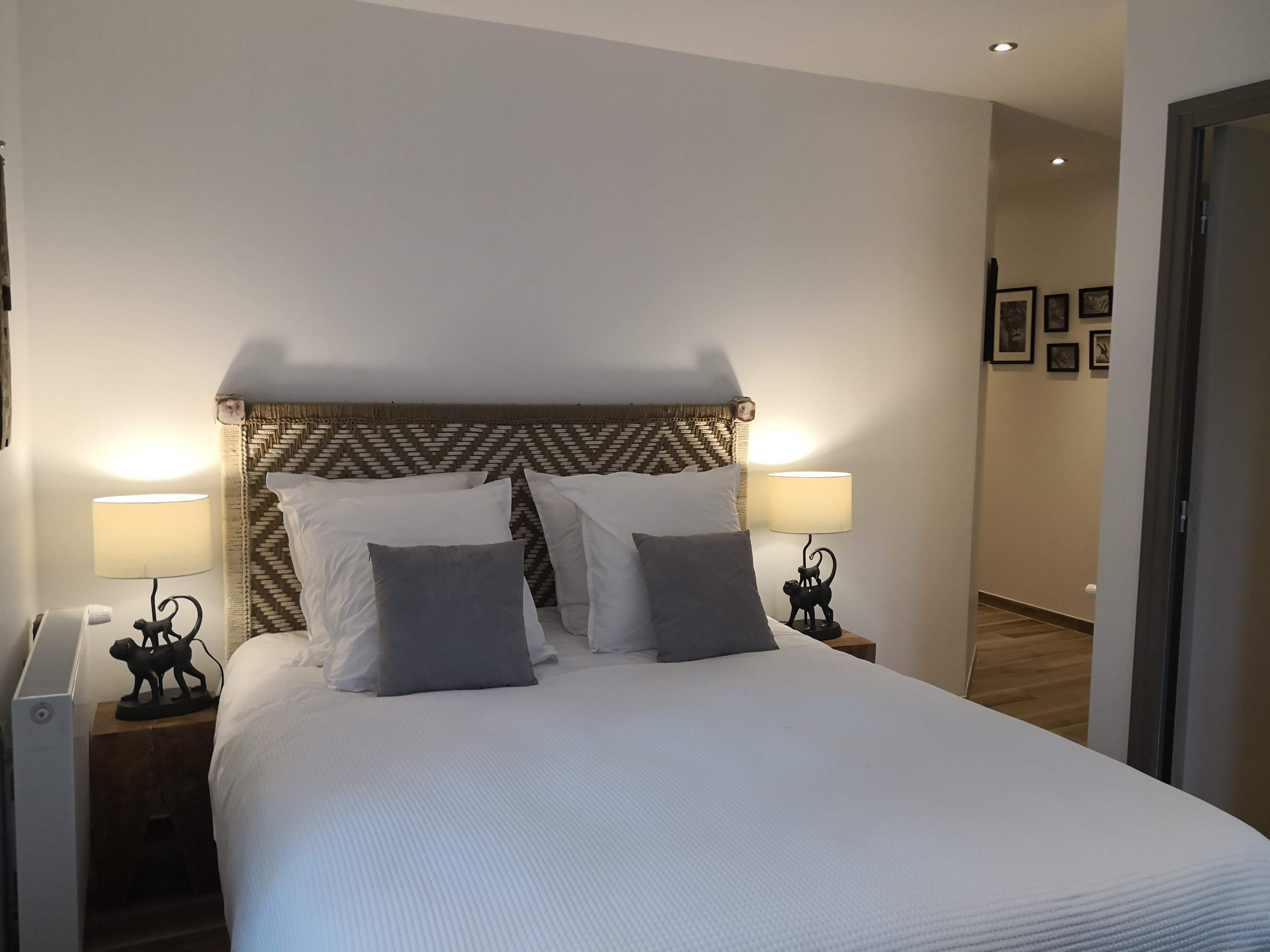 Grand lit avec tête de lit tressée en chevrons, coussins gris et blancs, lampes en forme de singes et vue vers un couloir avec cadres animaliers au mur.