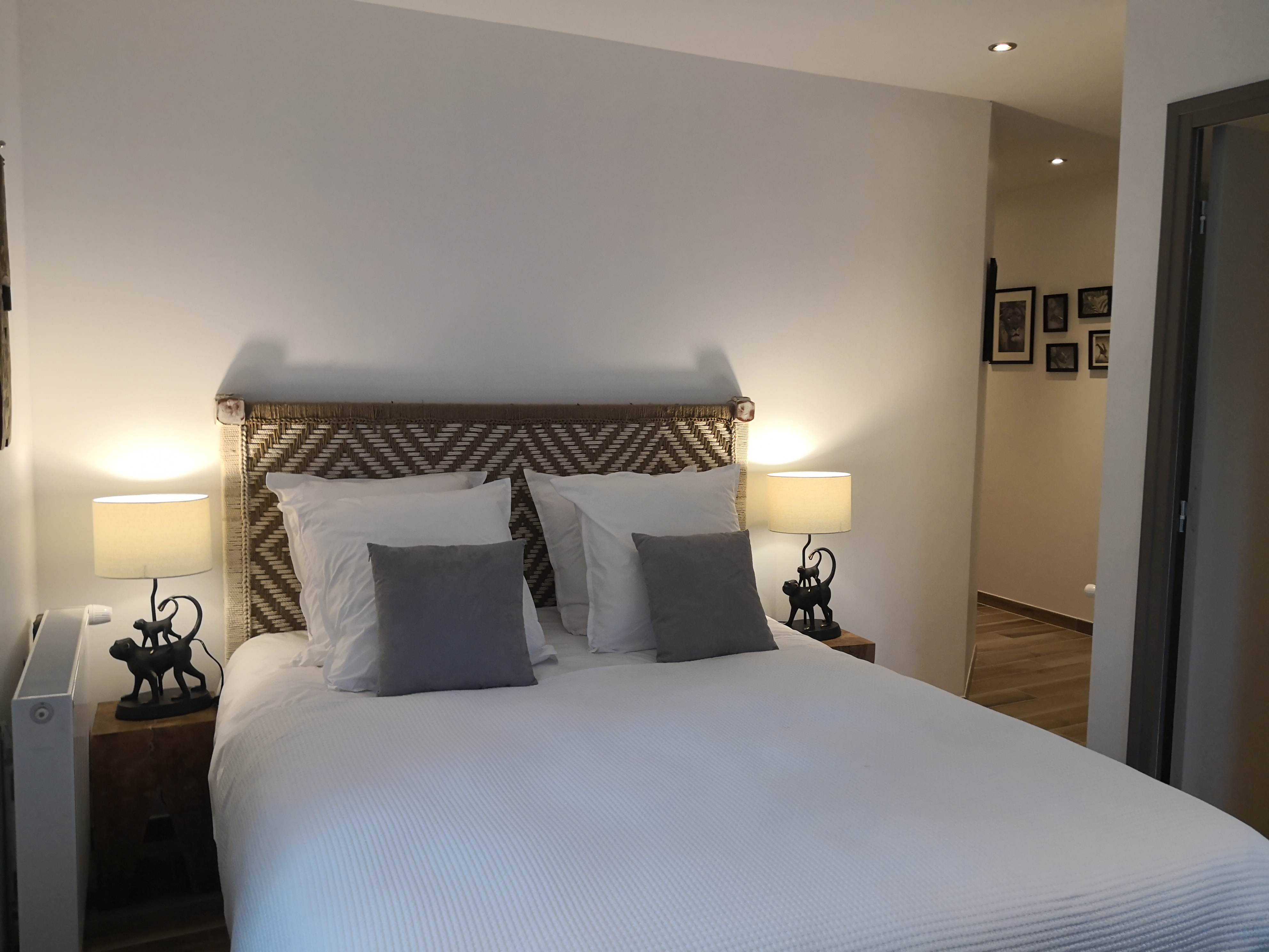 Grand lit avec tête de lit tressée en chevrons, coussins gris et blancs, lampes en forme de singes et vue vers un couloir avec cadres animaliers au mur.