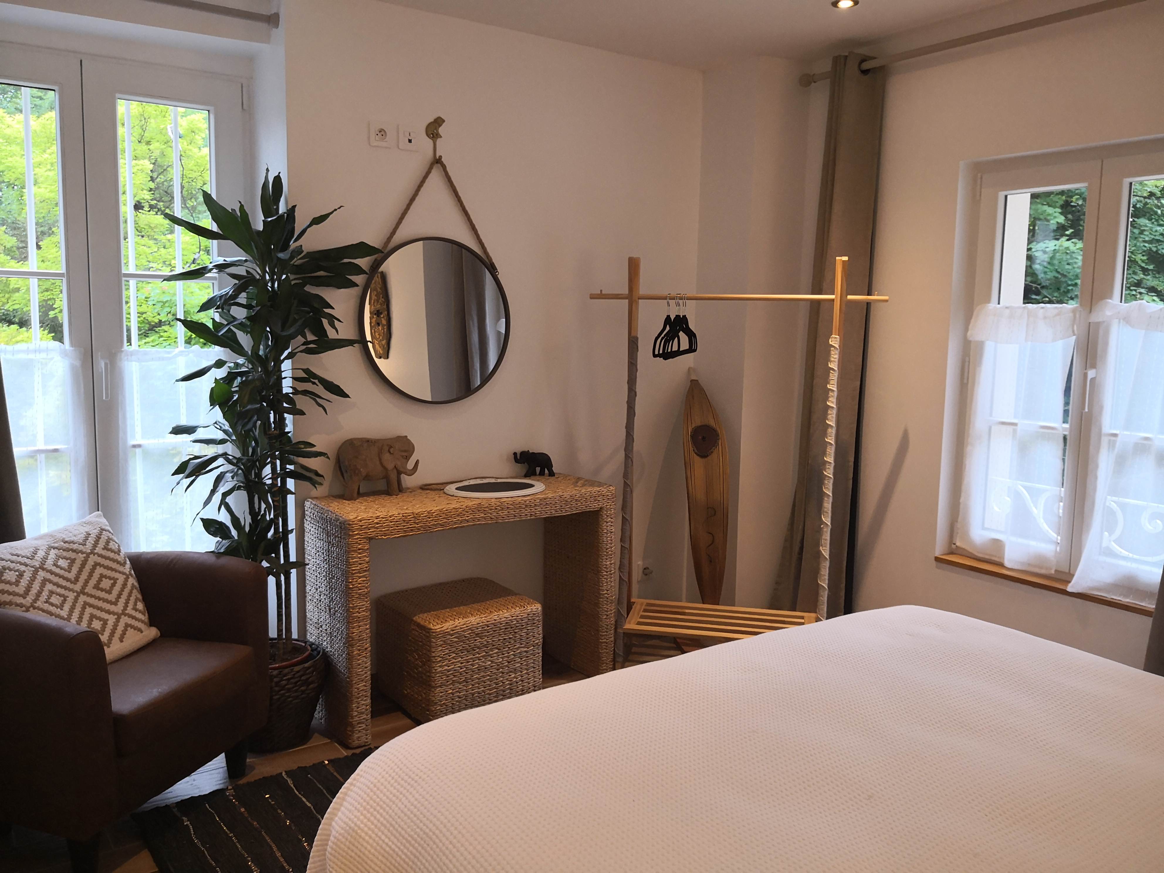Chambre avec miroir rond suspendu par une corde, console en fibres tressées, fauteuil en cuir, portant en bois avec planche de surf décorative et fenêtres donnant sur la verdure.