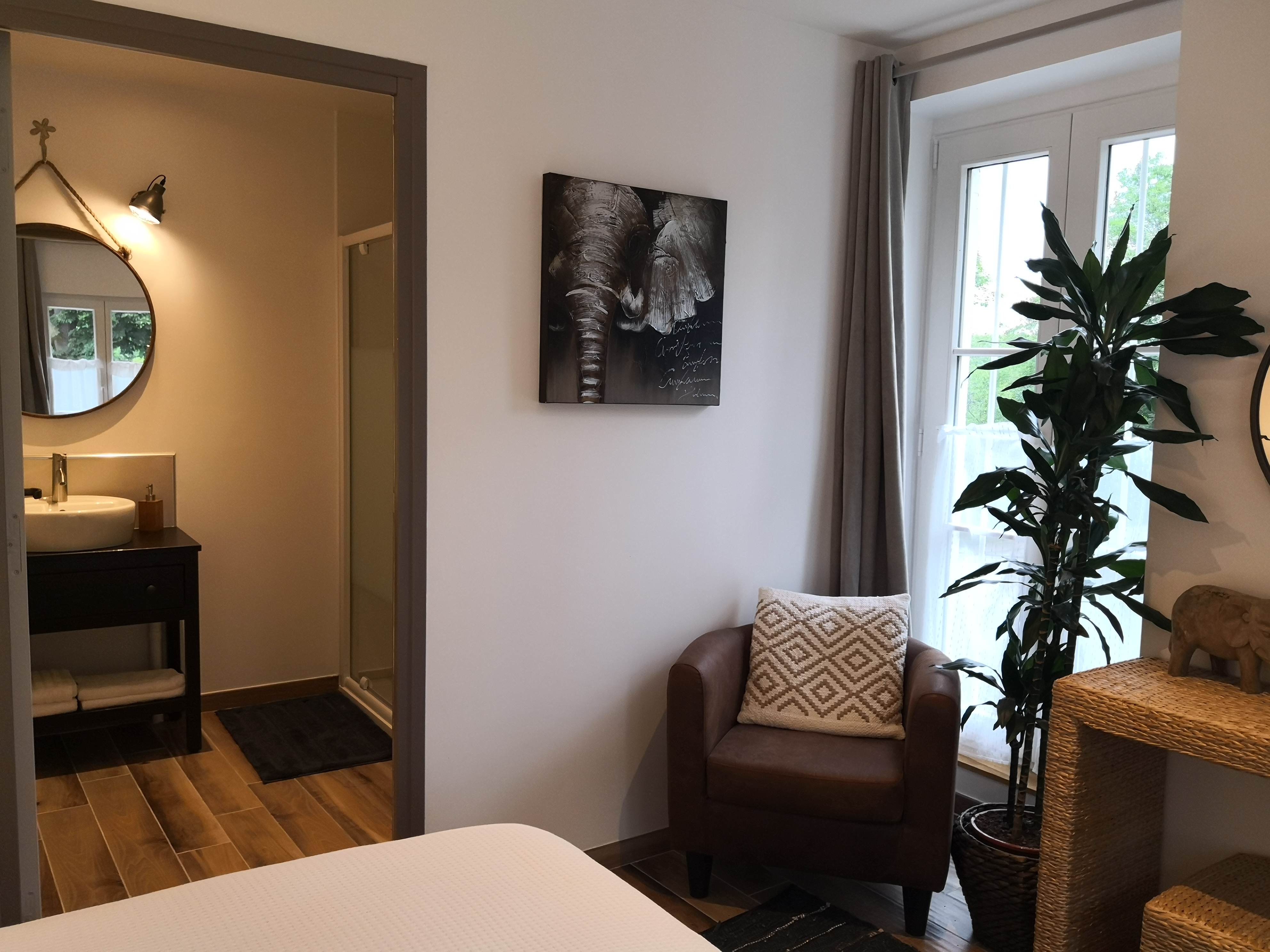 Coin chambre avec fauteuil en cuir brun, tableau d'éléphant, plante verte devant une fenêtre et vue sur la salle de bain avec vasque noire et miroir rond.